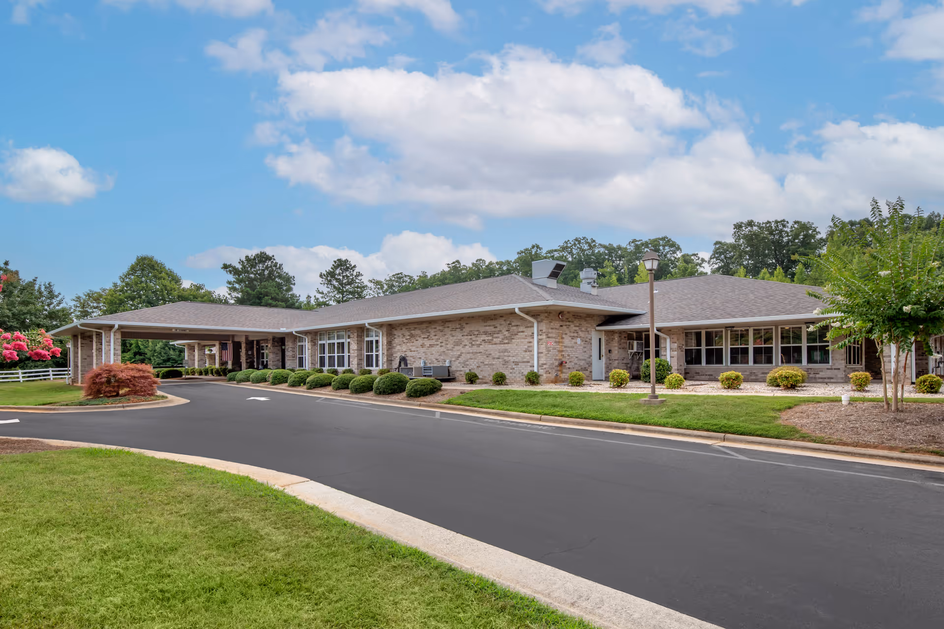 Exterior view of a single-story brick building with a covered entrance and a driveway in front, surrounded by well-maintained landscaping including bushes, trees, and grass under a partly cloudy sky.