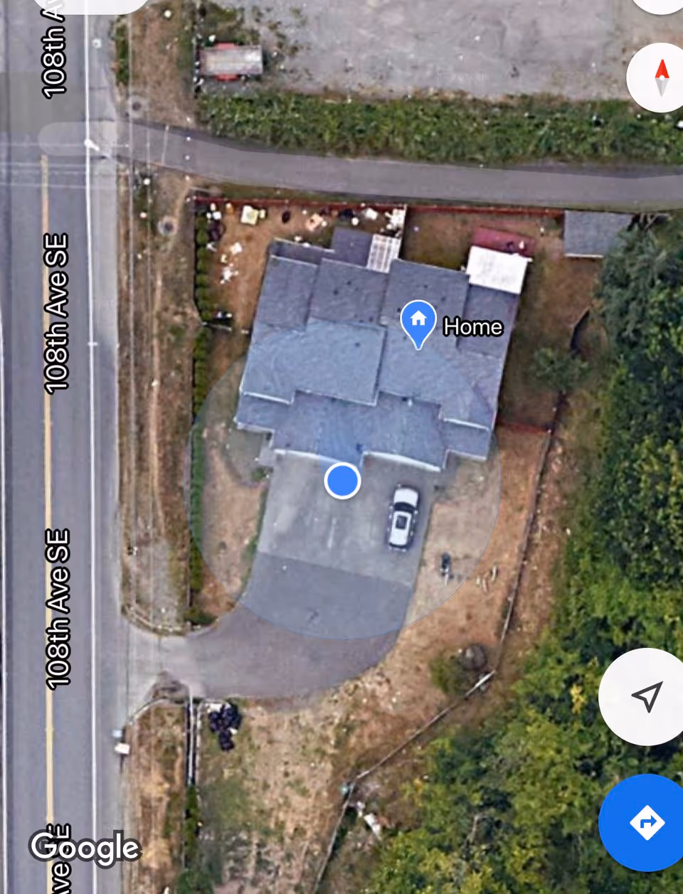 Aerial view of a residential property located at the corner of 108th Ave SE. The property includes a house with a driveway and a parked car. Surrounding the house are patches of grass, some trees, and a paved road.
