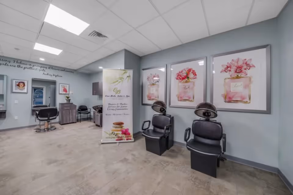 Interior view of a salon area in a senior living facility with two black hair drying chairs on the right, three framed floral perfume bottle artworks on the wall behind them, a banner stand with spa and beauty services information, and a styling chair with a mirror on the left side. The room has light gray walls and a tiled floor.