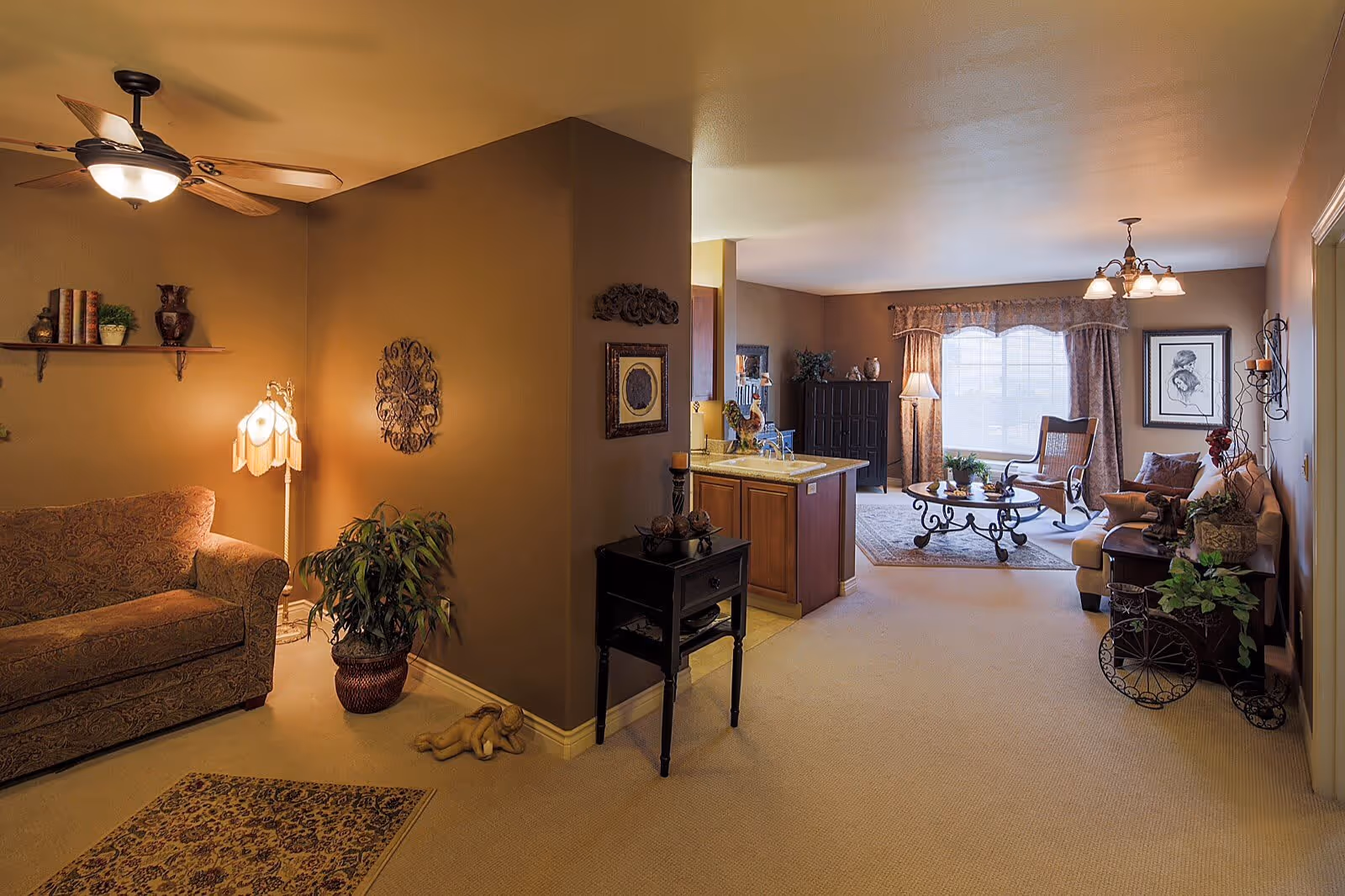 A cozy living room featuring a patterned sofa, a decorative lamp, and a small table with plants. The room has warm brown walls, a ceiling fan, and a large window with curtains, allowing natural light to fill the space.