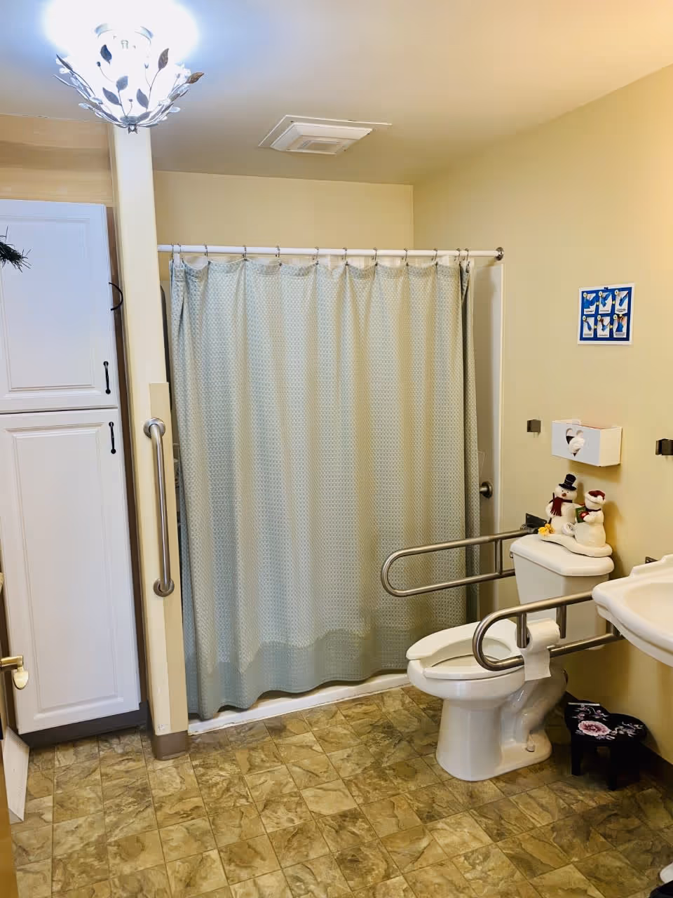 A senior-friendly bathroom with a toilet equipped with safety grab bars on both sides, a white sink, a shower with a light blue patterned curtain, a tall white cabinet, and a tiled floor. There are two small snowman decorations on top of the toilet tank and a small black step stool with a floral design near the sink.