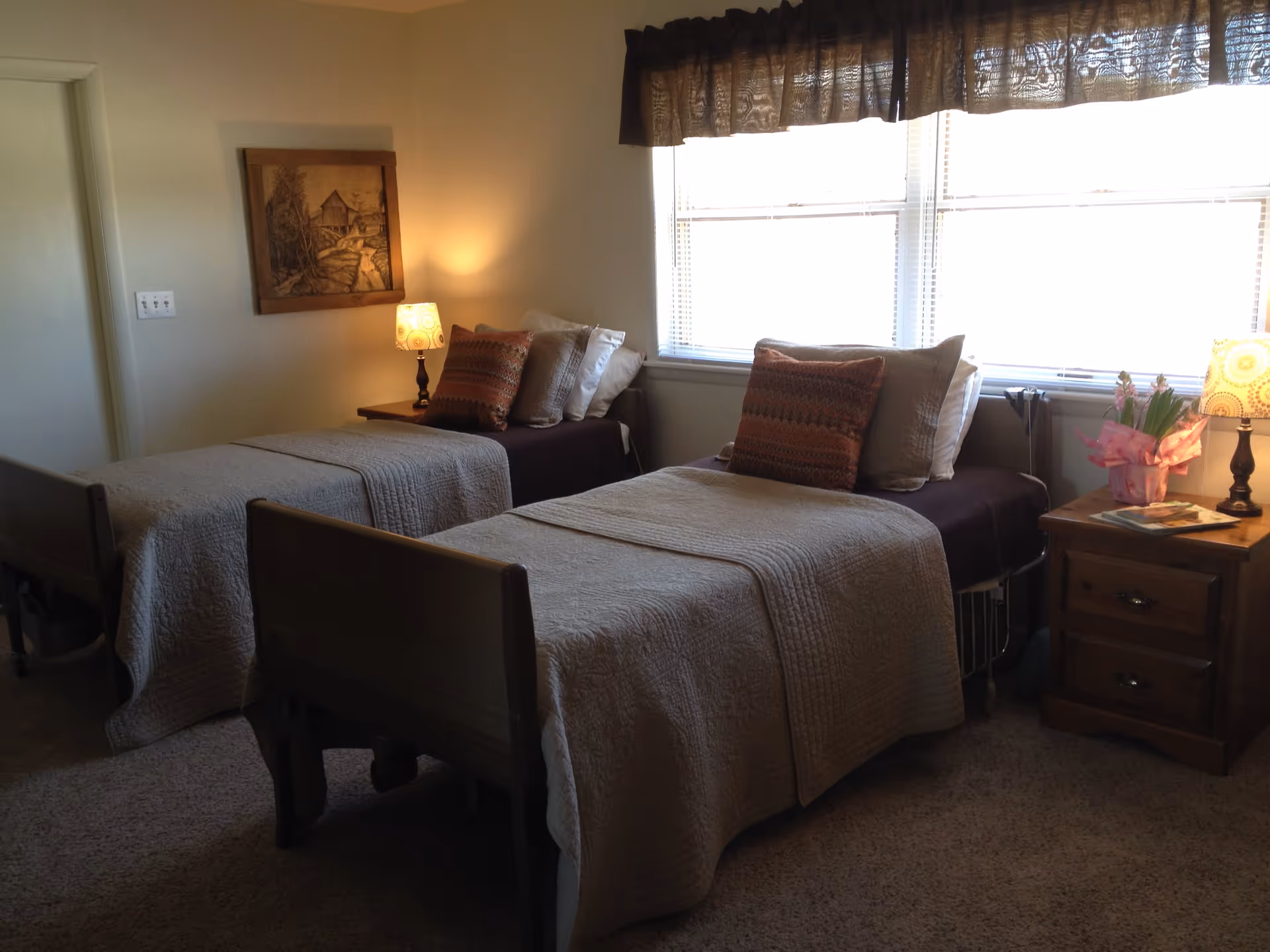 A cozy bedroom with two single beds, each covered with beige quilts and multiple pillows. There is a wooden nightstand between the beds with a lamp and a small potted plant. A framed artwork hangs on the wall above one bed, and a window with blinds and a valance lets in natural light.