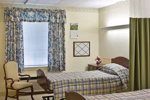 A small bedroom features two plaid beds, curtains, a chair, and a calendar on the wall.