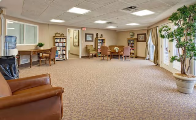 A spacious common area with carpeted floor, featuring a brown leather armchair in the foreground, a water cooler and a wooden table with chairs along the left wall, bookshelves filled with books, a round table with four chairs in the center, framed artwork on the walls, large windows with beige curtains on the right, and a large potted plant near the windows.