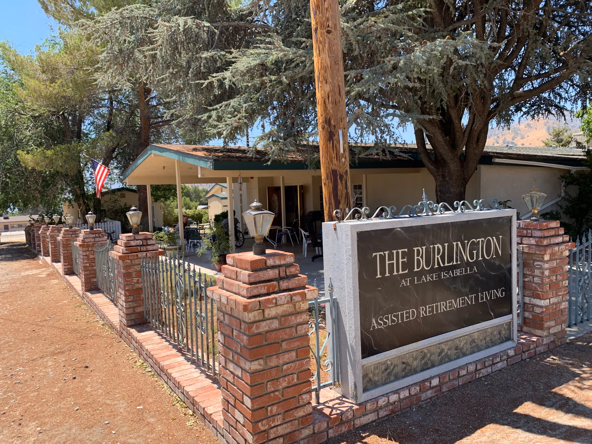 Exterior view of The Burlington Assisted Living facility showing a brick and metal fence with lamps, a large sign reading 'The Burlington at Lake Isabella Assisted Retirement Living,' a covered porch area with chairs, trees, and an American flag.