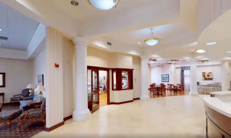 Interior view of a senior living facility featuring a spacious lobby area with large white columns, beige tiled flooring, and cream-colored walls with subtle vertical stripes. To the left, there is a seating area with upholstered chairs and a lamp on a side table. In the background, a dining area with wooden tables and chairs is visible, along with framed artwork on the walls and ceiling fans. The right side shows part of a curved reception desk.