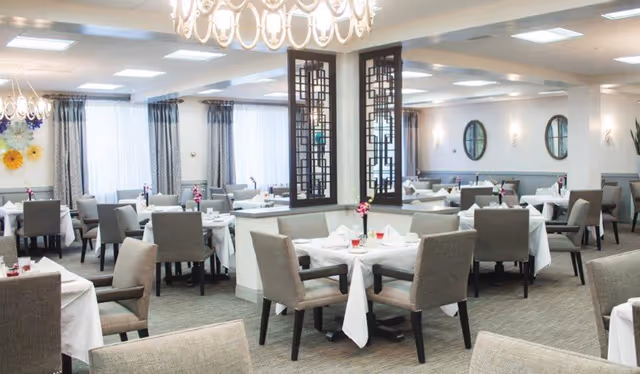 A bright and spacious dining room with multiple tables covered in white tablecloths, each set with glasses and small flower arrangements. The room features gray upholstered chairs, large windows with sheer curtains, decorative wall mirrors, and modern chandeliers hanging from the ceiling.
