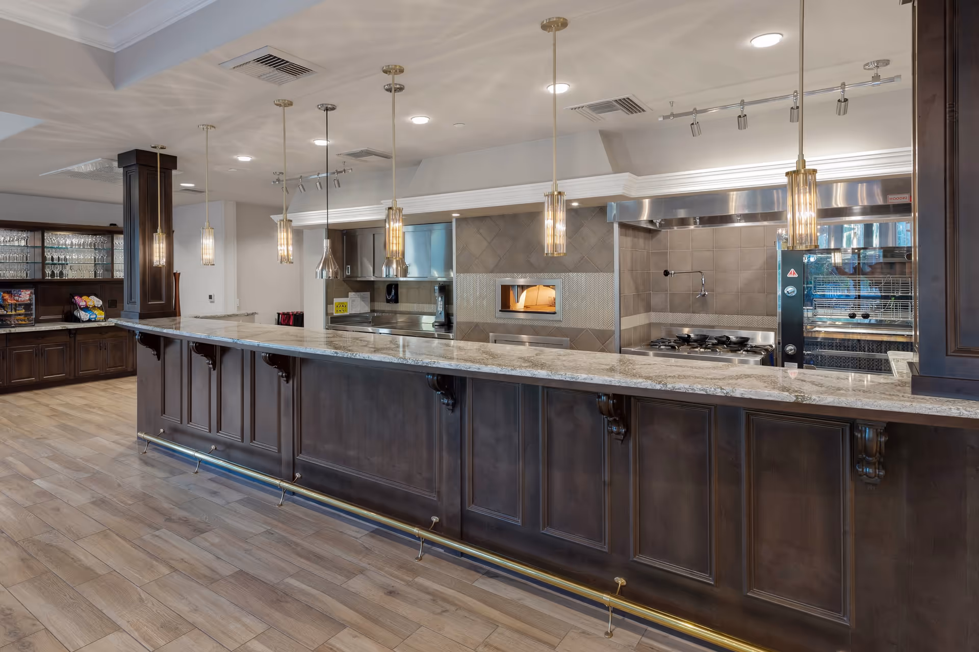 A modern kitchen area with a long marble countertop bar, dark wood cabinetry, pendant lights hanging from the ceiling, and stainless steel appliances including a stove and oven. There are shelves with glassware and snacks on the left side.
