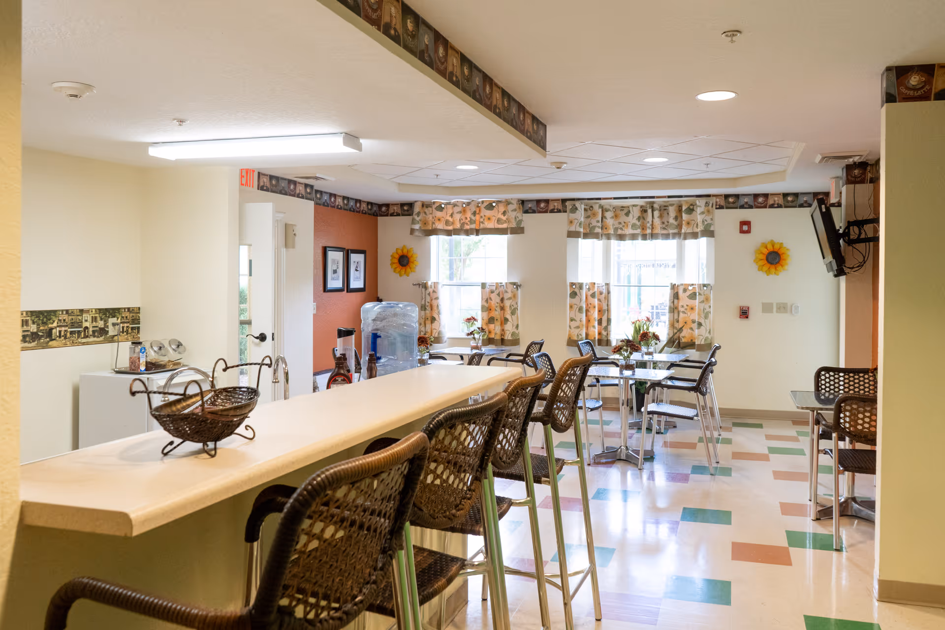 A bright dining area in a senior living facility with a long counter and several wicker bar stools. There are multiple small tables with chairs near windows adorned with floral curtains. The floor has a colorful checkered pattern, and the walls are decorated with sunflower-themed accents and framed pictures. A water dispenser and a wall-mounted television are also visible.