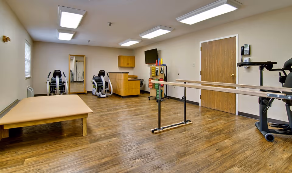 A rehabilitation or physical therapy room with wooden flooring, two exercise machines, a padded therapy table, parallel bars for walking exercises, a wall-mounted TV, a mirror, and a rack holding various exercise equipment. The room has beige walls, a wooden door, and fluorescent ceiling lights.