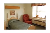 A small bedroom with a single bed covered in a green bedspread, a red upholstered armchair, a window with a valance, a wall-mounted air conditioning unit below the window, and a framed picture hanging on the wall.