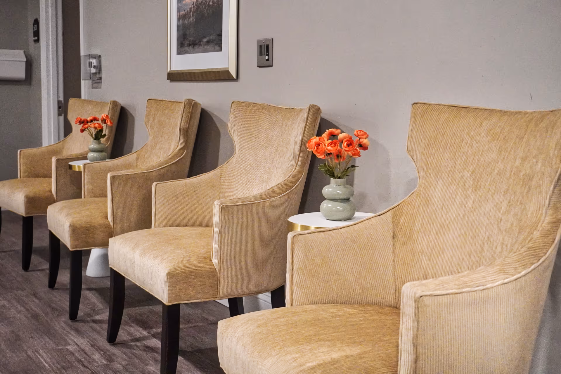 Row of beige upholstered armchairs with small side tables and vases of orange flowers along an indoor wall.