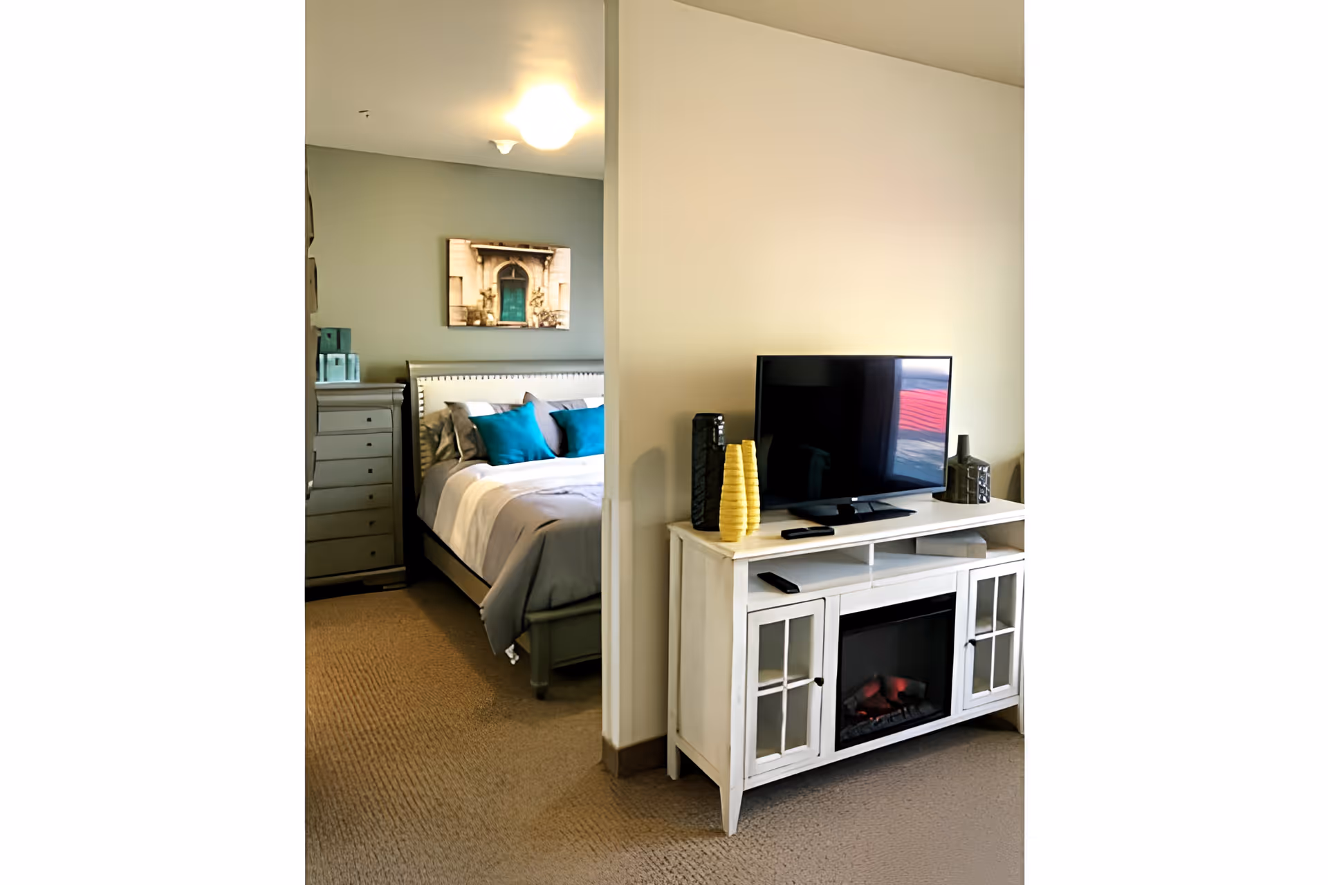 View of a cozy bedroom partially visible through an open doorway, featuring a bed with gray and teal pillows, a dresser, and a framed picture on the wall. In the foreground, there is a white TV stand with a flat-screen television, decorative vases, and an electric fireplace insert.