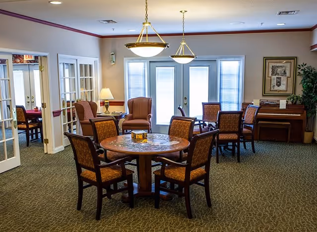 A well-lit common area in a senior living facility featuring a round wooden table with six upholstered chairs around it. In the background, there are two armchairs with a small table and lamp between them, a piano with a plant on top, and large windows with blinds letting in natural light. The room has carpeted flooring and neutral-colored walls with crown molding.
