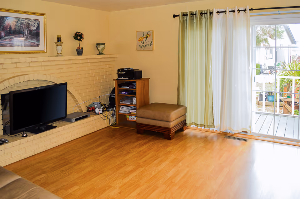 A living room with light wood flooring and beige walls. There is a built-in brick fireplace painted cream with a flat-screen TV placed inside the fireplace opening. On the mantel are decorative items including a framed landscape painting, a candle holder, a small potted plant, and a vase. Next to the fireplace is a small wooden shelving unit holding a printer and various items. In front of the shelving unit is a brown cushioned ottoman. To the right, there is a large sliding glass door with green and white curtains, leading to an outdoor area with plants and a deck.