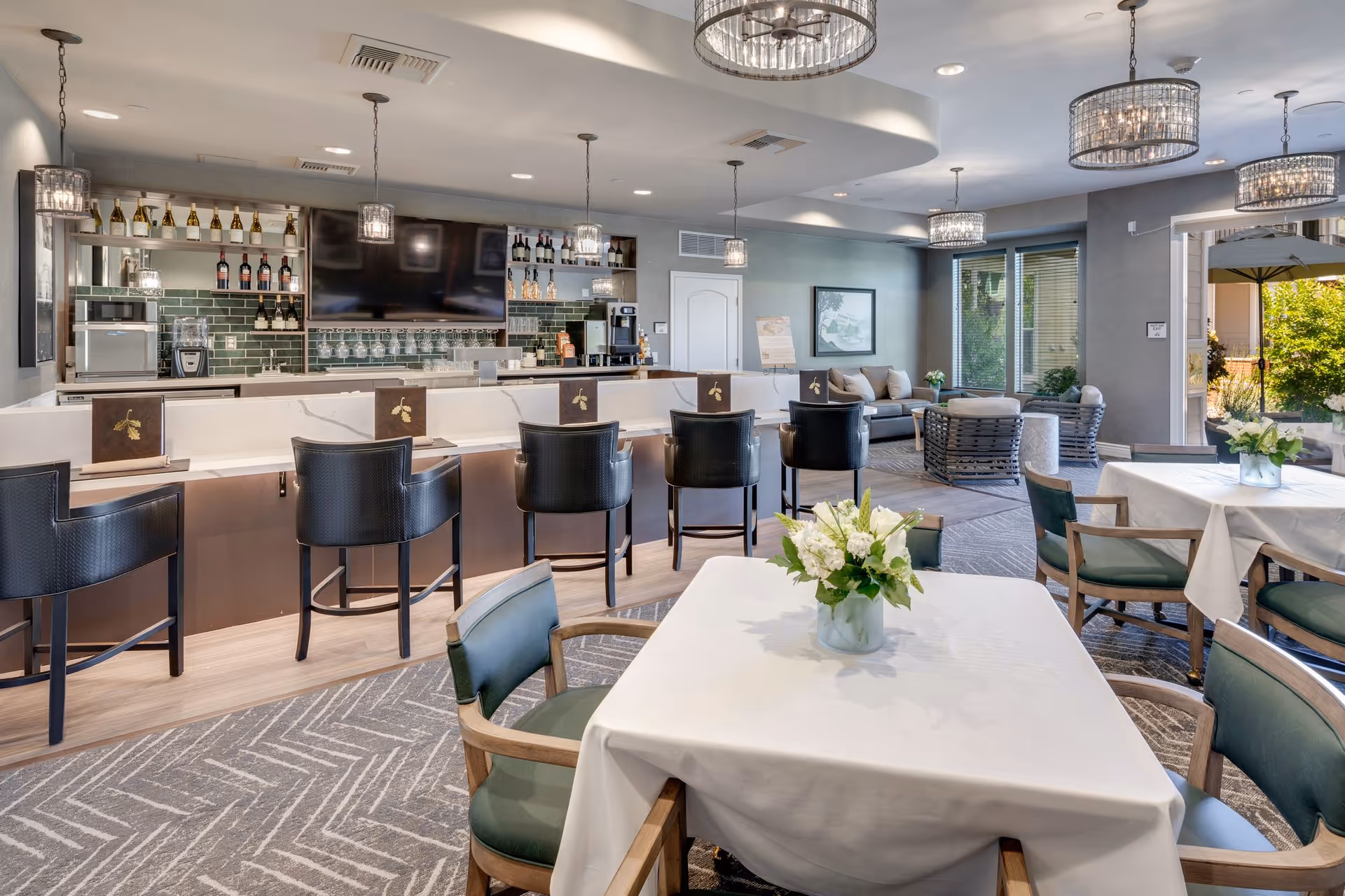 Bright dining and lounge area with tablecloth-covered tables, chairs, a bar with stools, pendant lighting, and a seating area by windows.