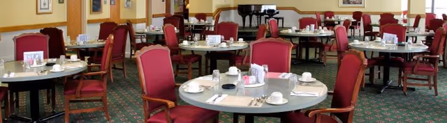 Dining room with round tables set with cups, saucers, utensils, and red upholstered chairs.