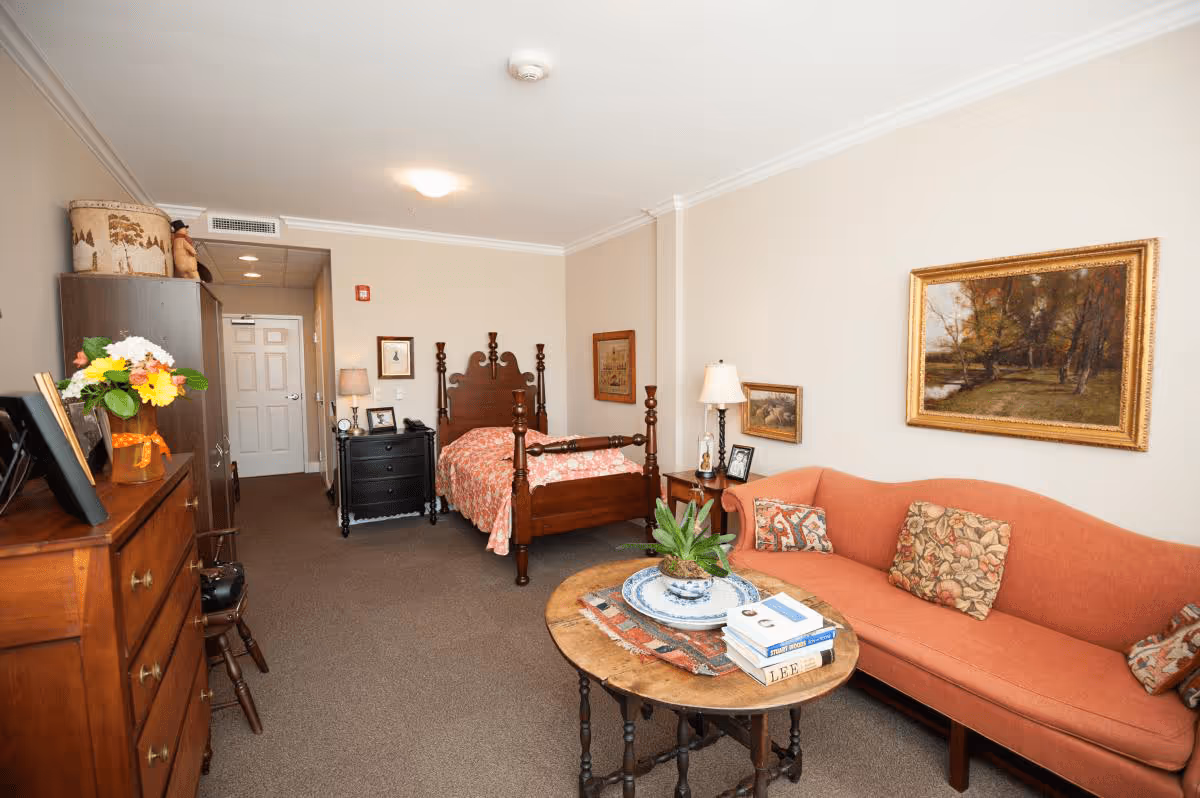A cozy room with a wooden four-poster bed covered with a floral bedspread, a coral-colored sofa with patterned cushions, a round wooden coffee table with books and a plant, a wooden dresser with a vase of flowers, and framed paintings on the beige walls.