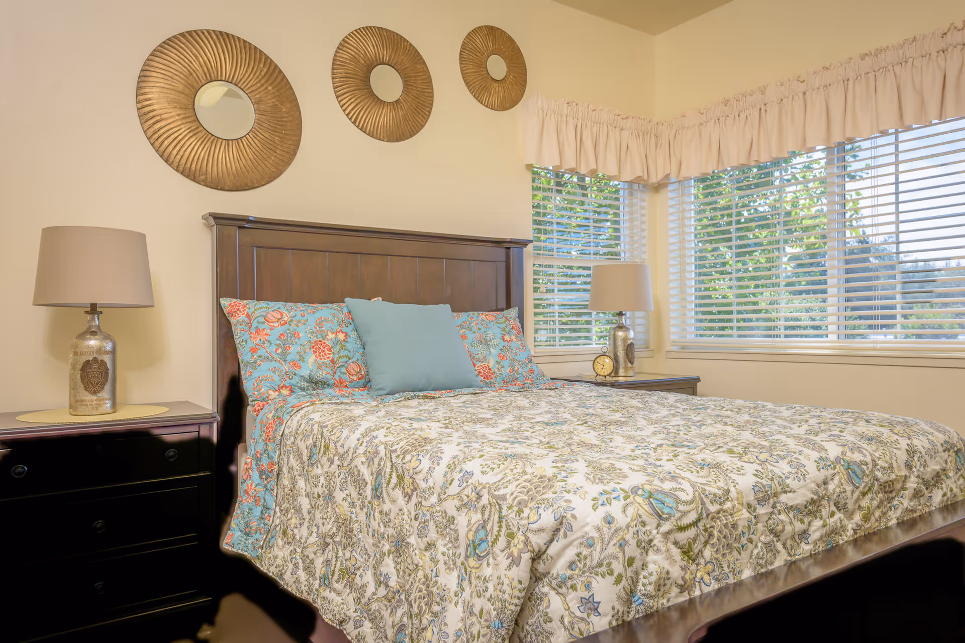 A bedroom with a wooden bed frame and a floral patterned bedspread in shades of blue, green, and beige. There are two matching floral pillows and one solid blue pillow on the bed. On either side of the bed are dark wooden nightstands with lamps featuring beige shades and decorative bases. Above the bed are three round, gold decorative wall mirrors. The room has large windows with white blinds and beige curtains, allowing natural light to brighten the space.