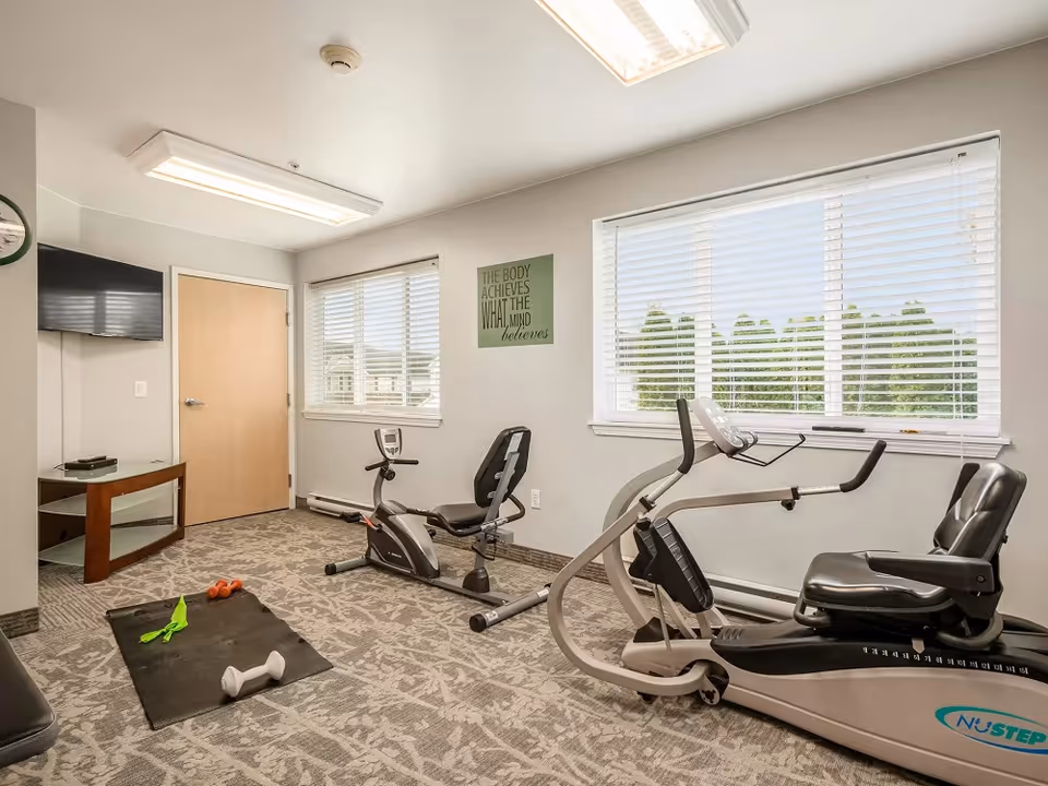A small exercise room with two recumbent stationary bikes, a black exercise mat with dumbbells and resistance bands on the floor, a wall-mounted TV, and two large windows with white blinds letting in natural light. A motivational sign on the wall reads 'The body achieves what the mind believes.'