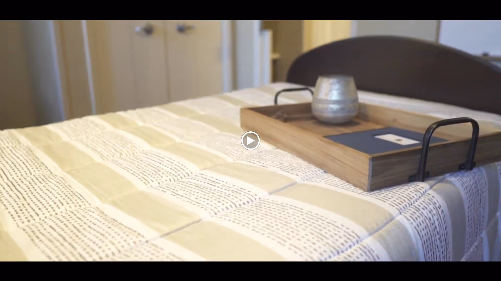 Close-up view of a bed with a beige and white patterned bedspread. On the bed is a wooden tray holding a metallic cup and a small book or card. In the background, there is a closed closet with double doors.