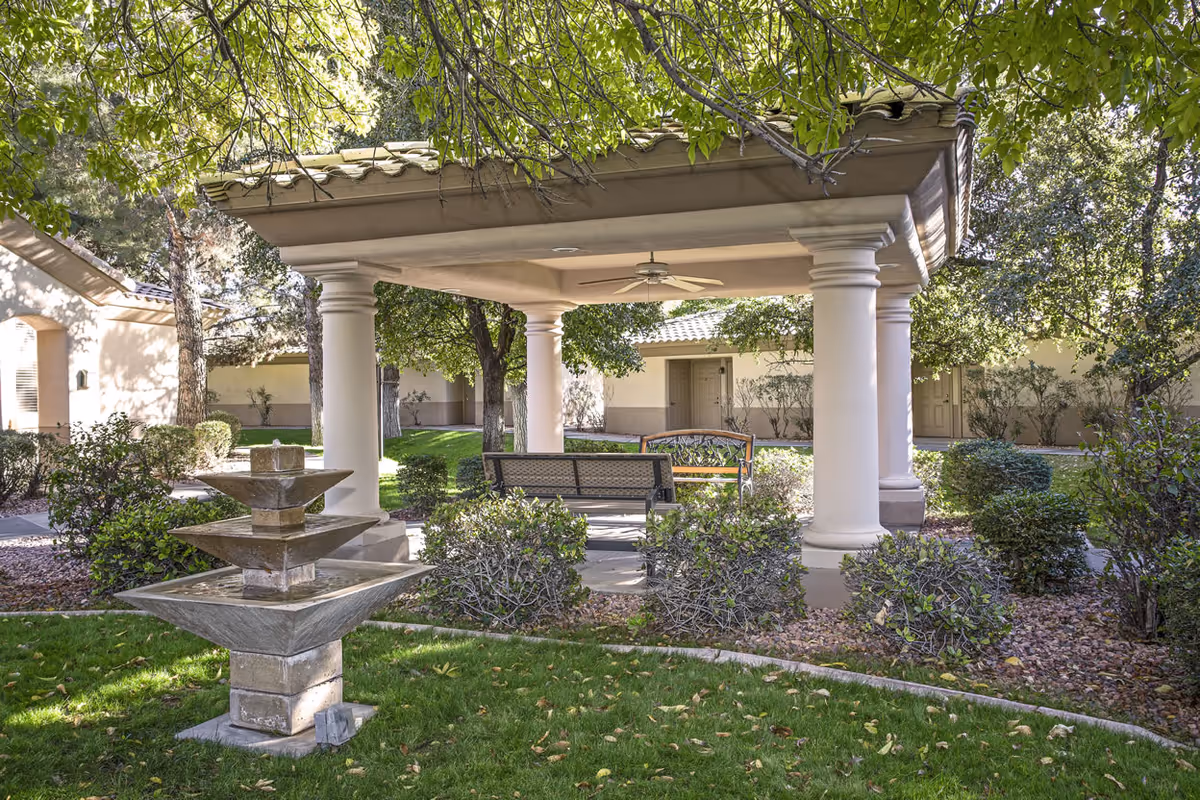 A shaded courtyard with a covered pavilion, benches, and a tiered fountain surrounded by trees and shrubs.