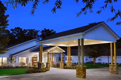 Front entrance of a senior living building at dusk with a lit porte-cochere supported by stone columns and landscaped grounds.