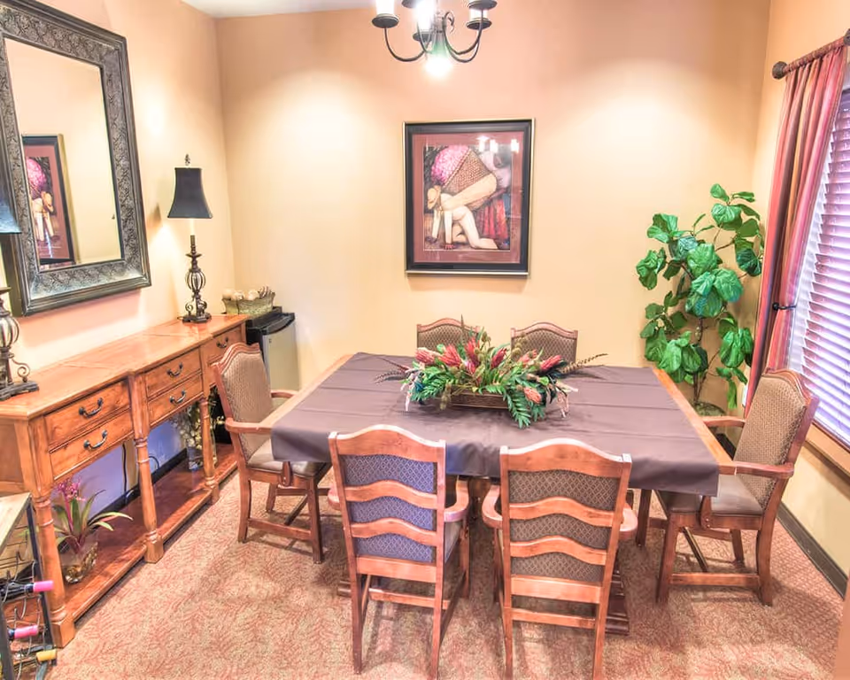 Dining room with a rectangular table covered by a tablecloth, six wooden chairs, a floral centerpiece, a sideboard with lamps, wall art and a potted plant.