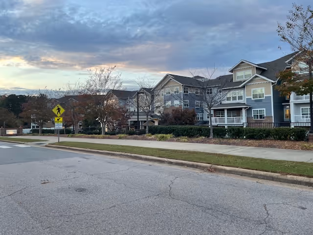 Exterior view of a multi-story residential building with blue and white siding, surrounded by trees and bushes, under a cloudy sky at dusk. A pedestrian crossing sign is visible near the sidewalk along a paved road.