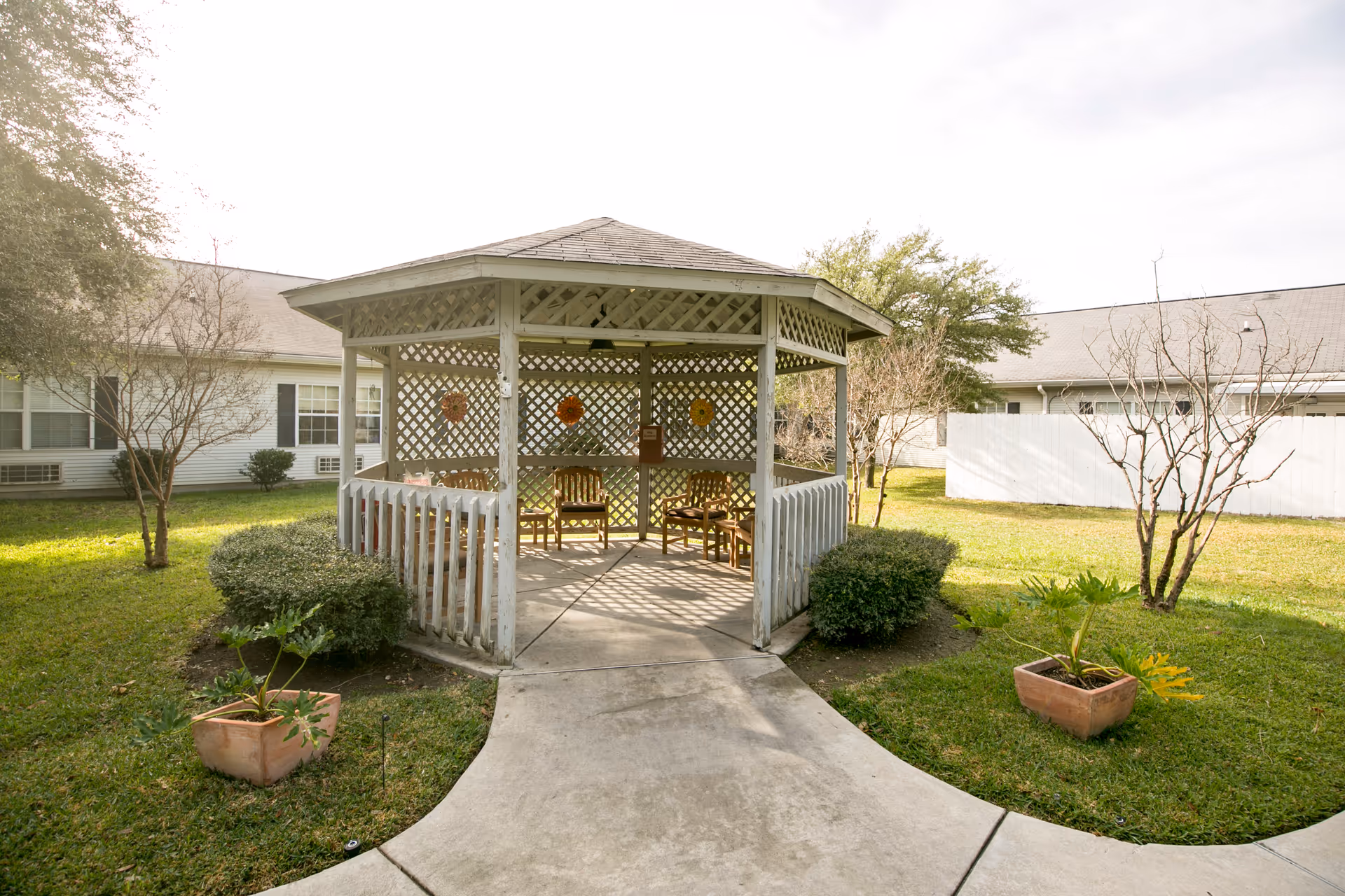 A white wooden gazebo with lattice walls and a shingled roof sits in a grassy courtyard surrounded by bushes and small trees. Inside the gazebo are several wooden chairs arranged around the perimeter. The courtyard is part of a senior living facility with single-story buildings visible in the background.
