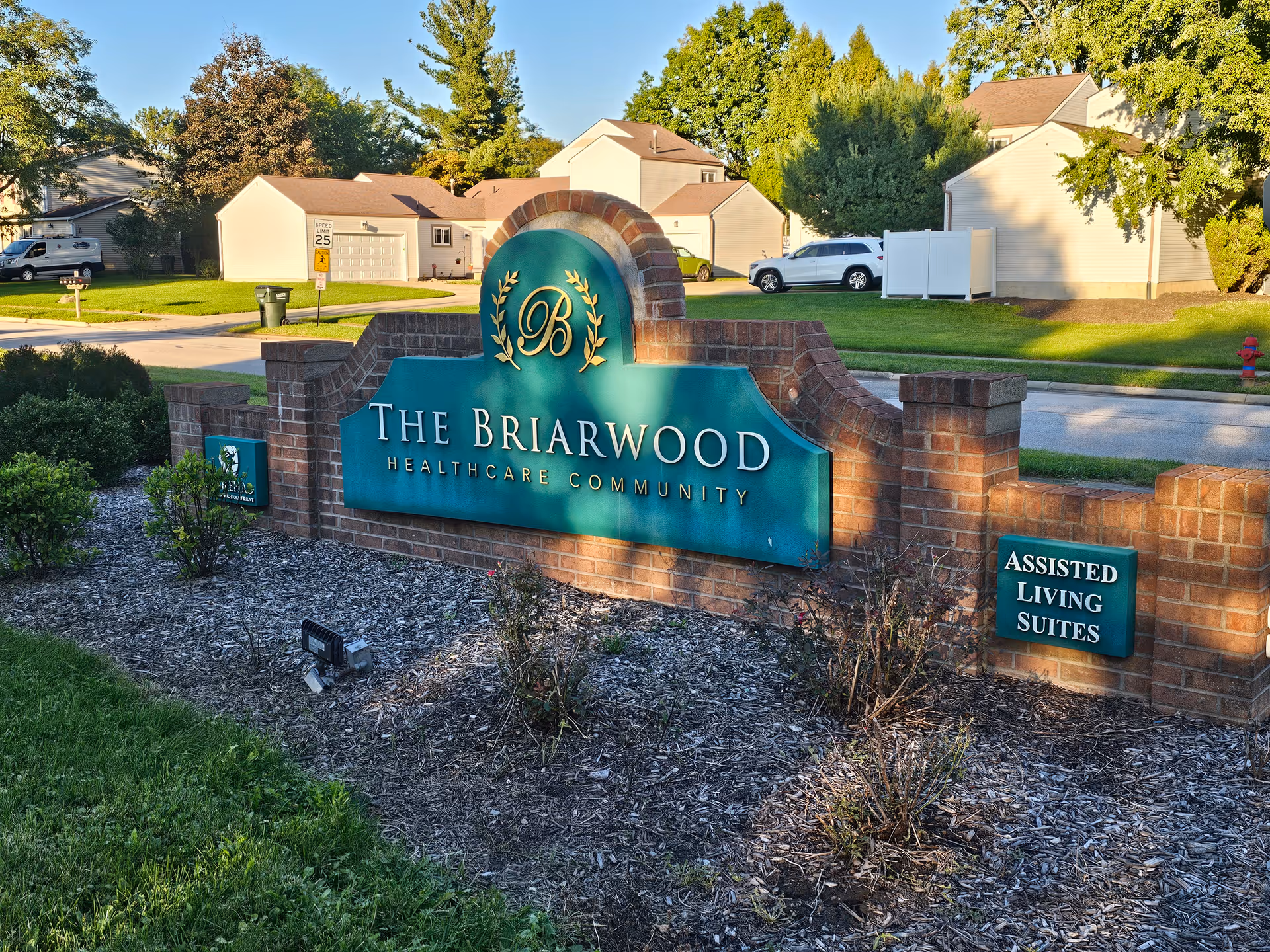 Brick entrance sign reading 'The Briarwood Healthcare Community' with landscaping and neighborhood homes in the background.