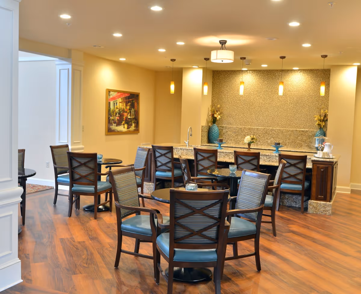 A well-lit dining area with several round tables and wooden chairs with blue cushions. In the background, there is a granite countertop bar with high chairs, decorative vases, and pendant lights hanging from the ceiling. The floor is wooden, and a framed painting is visible on the wall.