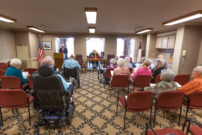 A group of elderly people seated in a room facing a man standing at a podium. The room has patterned carpet, an American flag, and a Christian flag. Some attendees are in wheelchairs, and the setting appears to be a community or meeting room in a senior living facility.