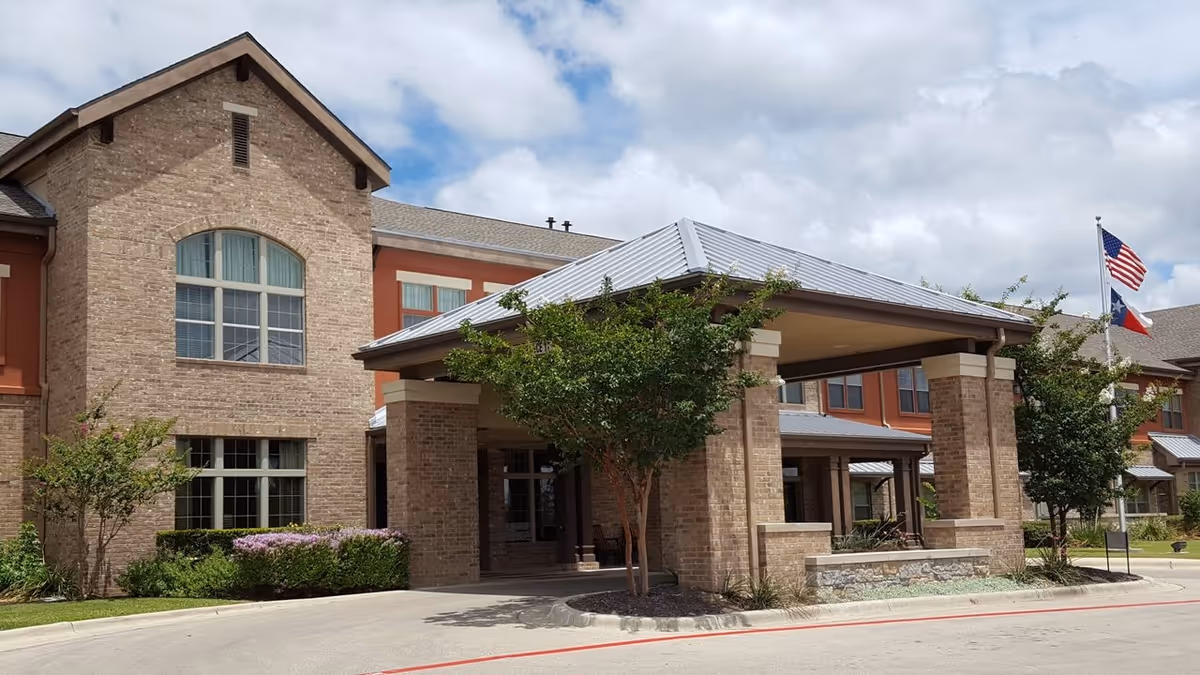Exterior view of The Wesleyan Assisted Living and Memory Care building showing a brick facade with large windows, a covered entrance supported by brick columns, landscaping with trees and shrubs, and American and Texas flags on flagpoles under a partly cloudy sky.
