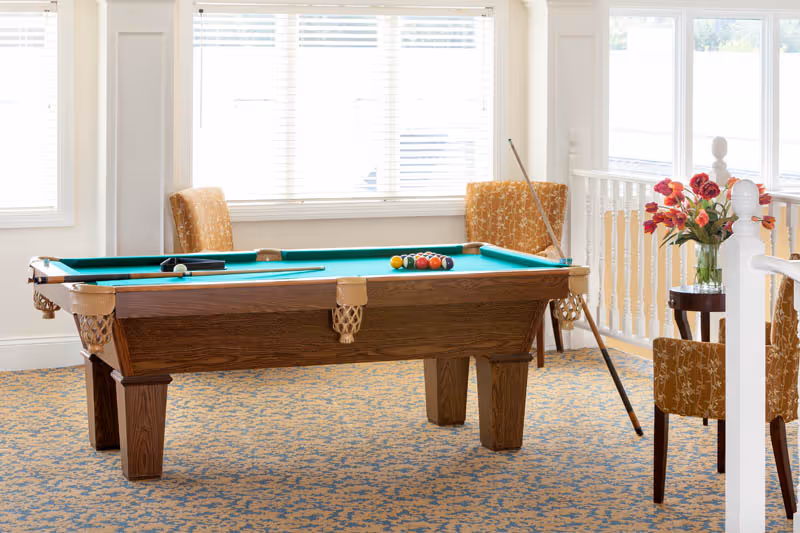A pool table in a bright common room with upholstered chairs, patterned carpet, and windows.