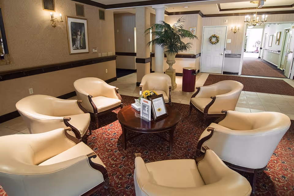 A cozy seating area in a senior living facility with six cream-colored armchairs arranged in a circle around a round wooden table. The table has framed information and a small flower arrangement. The room features a patterned red carpet, beige walls with sconces, a large potted plant, and a hallway leading to other rooms.