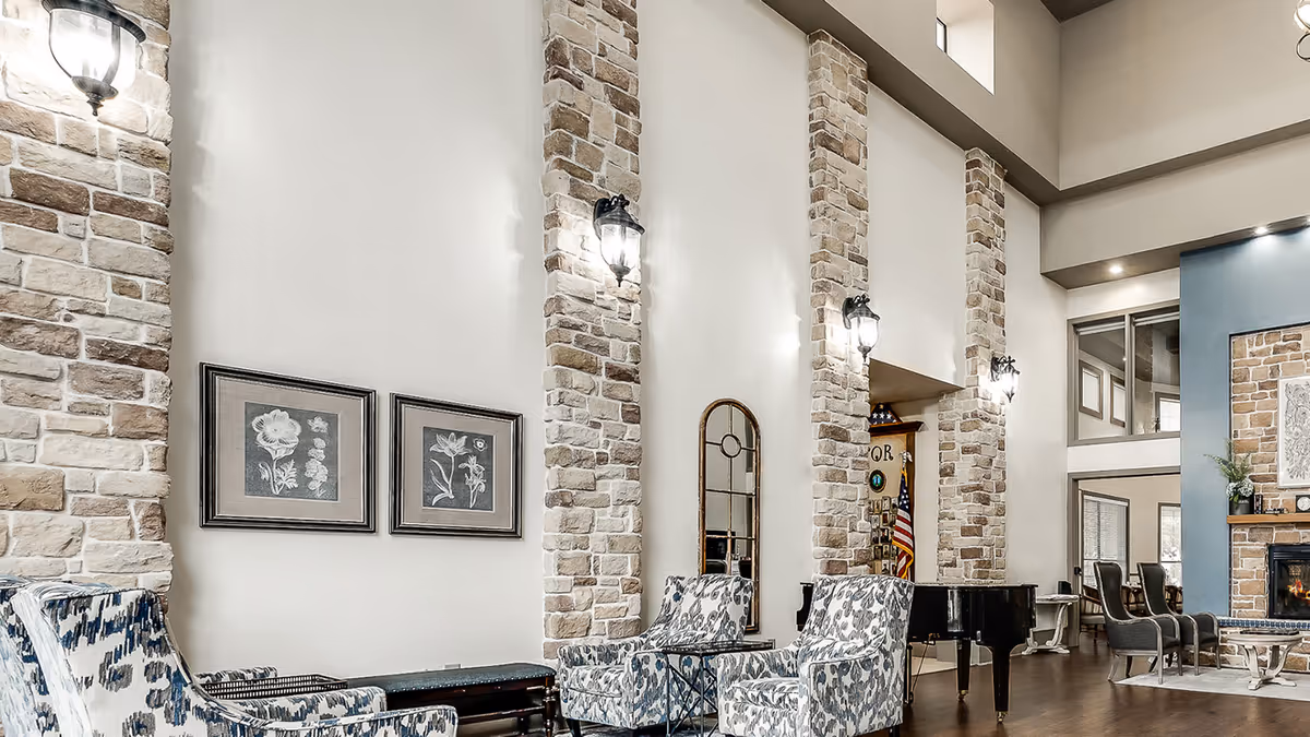 High-ceilinged communal living area with stone columns, patterned armchairs, a grand piano, and a fireplace.