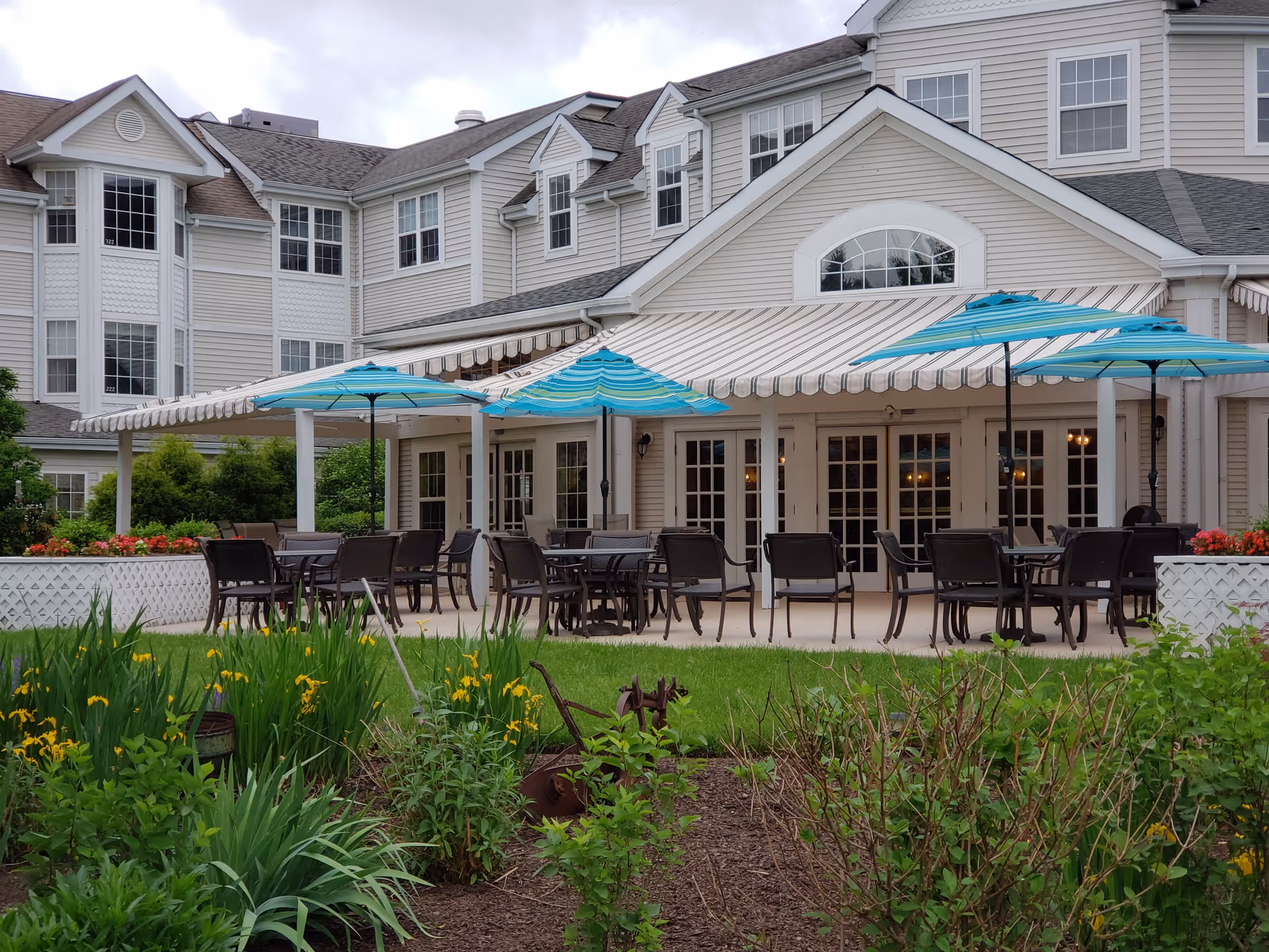 Outdoor patio area with tables and blue-striped umbrellas in front of a multi-story senior living building.
