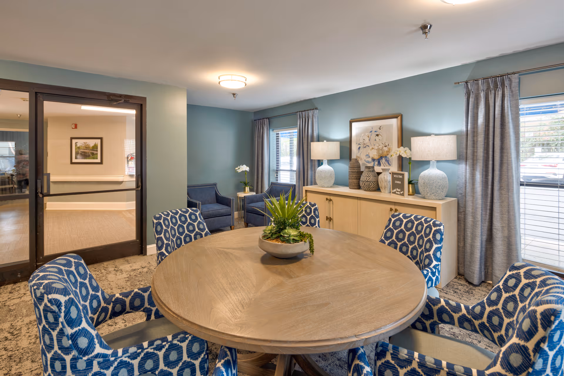 Round wooden table surrounded by patterned blue chairs in a well-lit communal dining/lounge room with a sideboard, lamps, and windows.