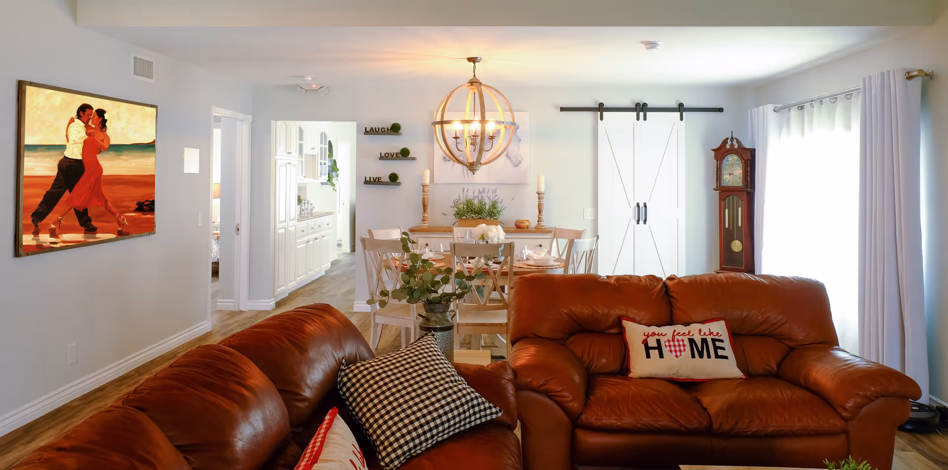 Open-plan living and dining area with brown leather sofas in the foreground, a dining table under a chandelier, and white sliding barn doors in the background.