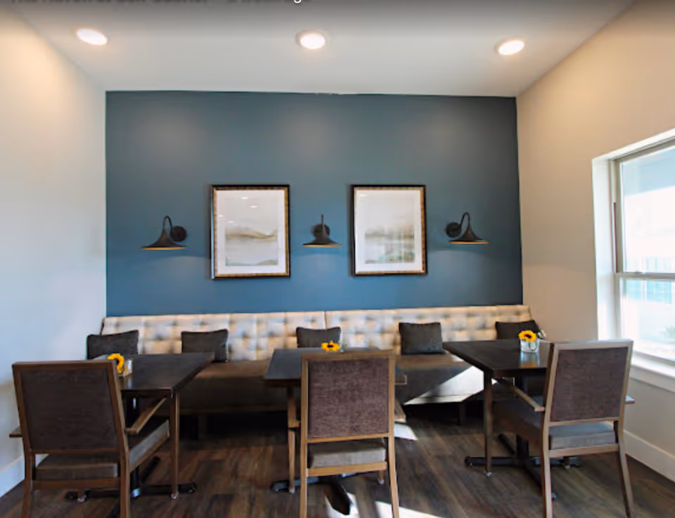 A cozy dining area with a long tufted bench against a blue accent wall, two framed pictures, and three wall-mounted lamps. There are four wooden chairs around two dark wooden tables, each table decorated with a small vase containing yellow flowers. A large window on the right lets in natural light.