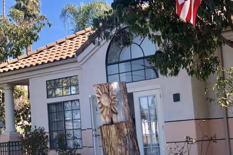 Front entrance of a light-colored residential care building with an arched window, French doors, a carved wooden sculpture, and an American flag.