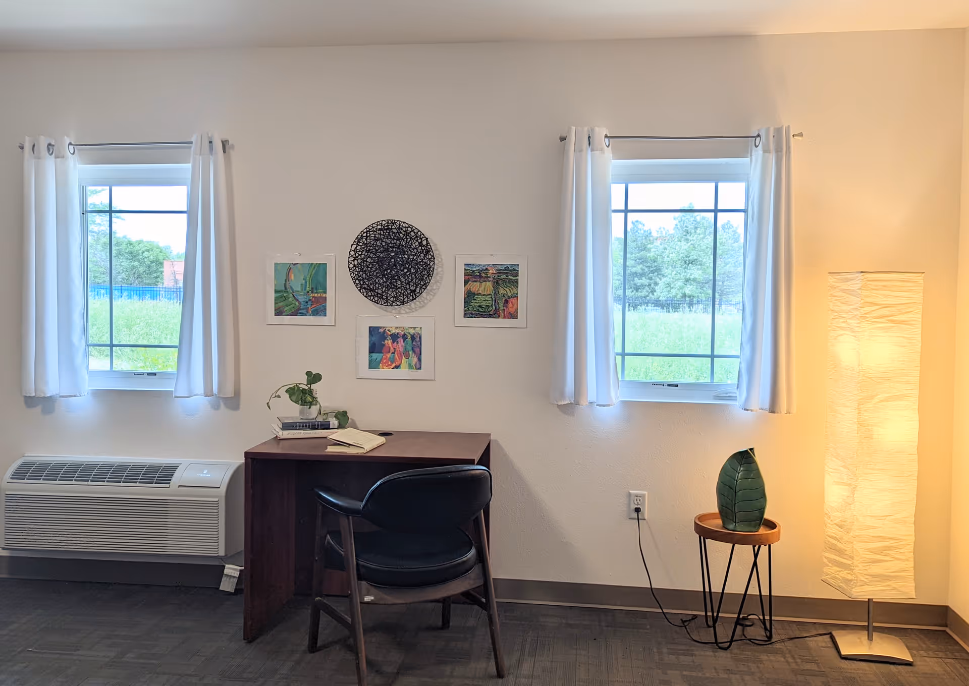 A small workspace area in a room with two windows covered by white curtains. Between the windows is a wooden desk with a black chair. On the desk are a small plant, a book, and a notebook. Above the desk are four pieces of wall art, including a circular black design and three colorful paintings. To the right of the desk is a small round side table with a green leaf decoration and a tall floor lamp with a white textured shade. The room has light-colored walls and dark flooring.