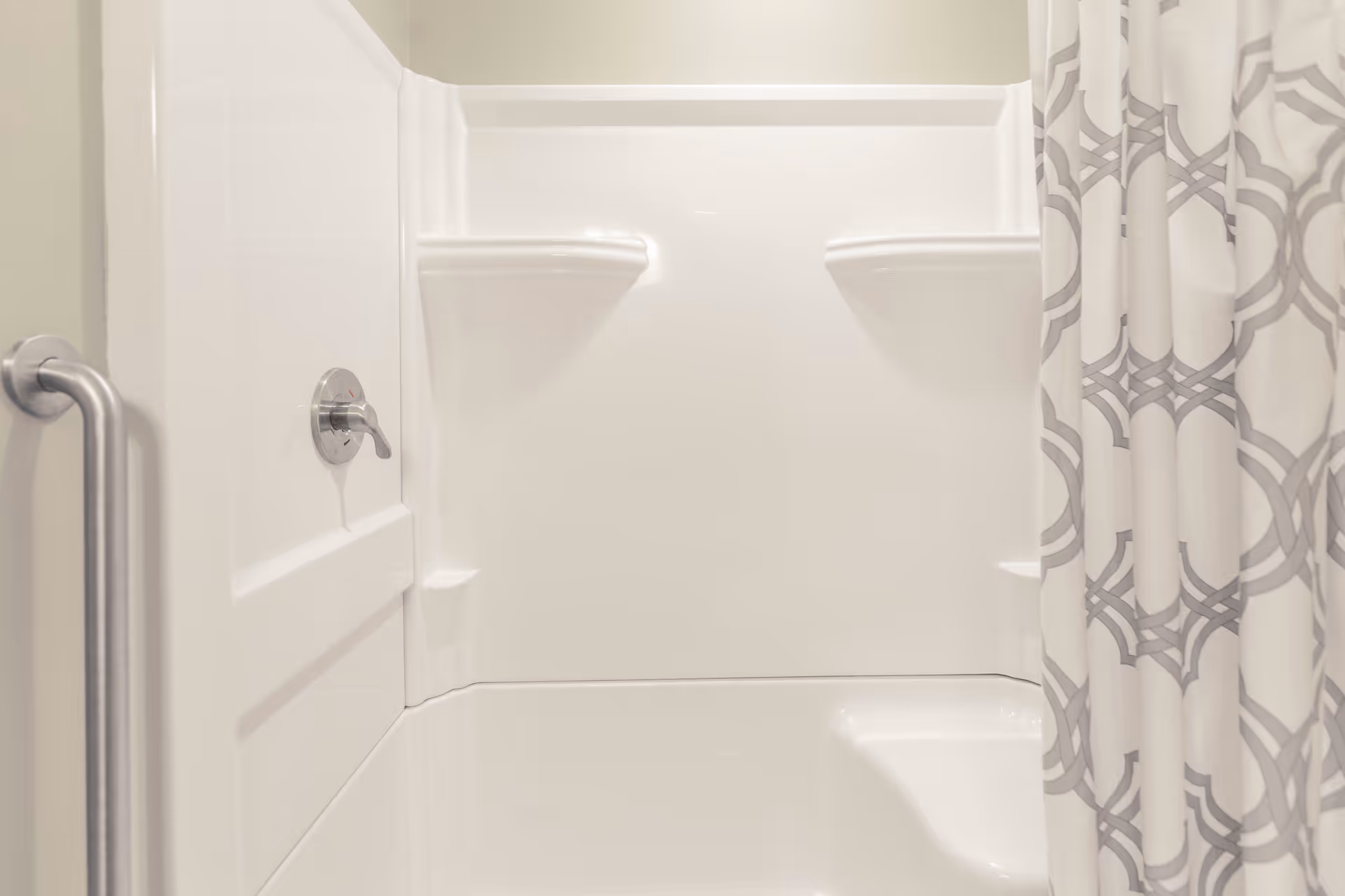 A clean white bathtub with built-in shelves and a silver faucet handle. A patterned shower curtain with gray geometric designs is partially visible on the right side. A silver grab bar is mounted on the left wall.