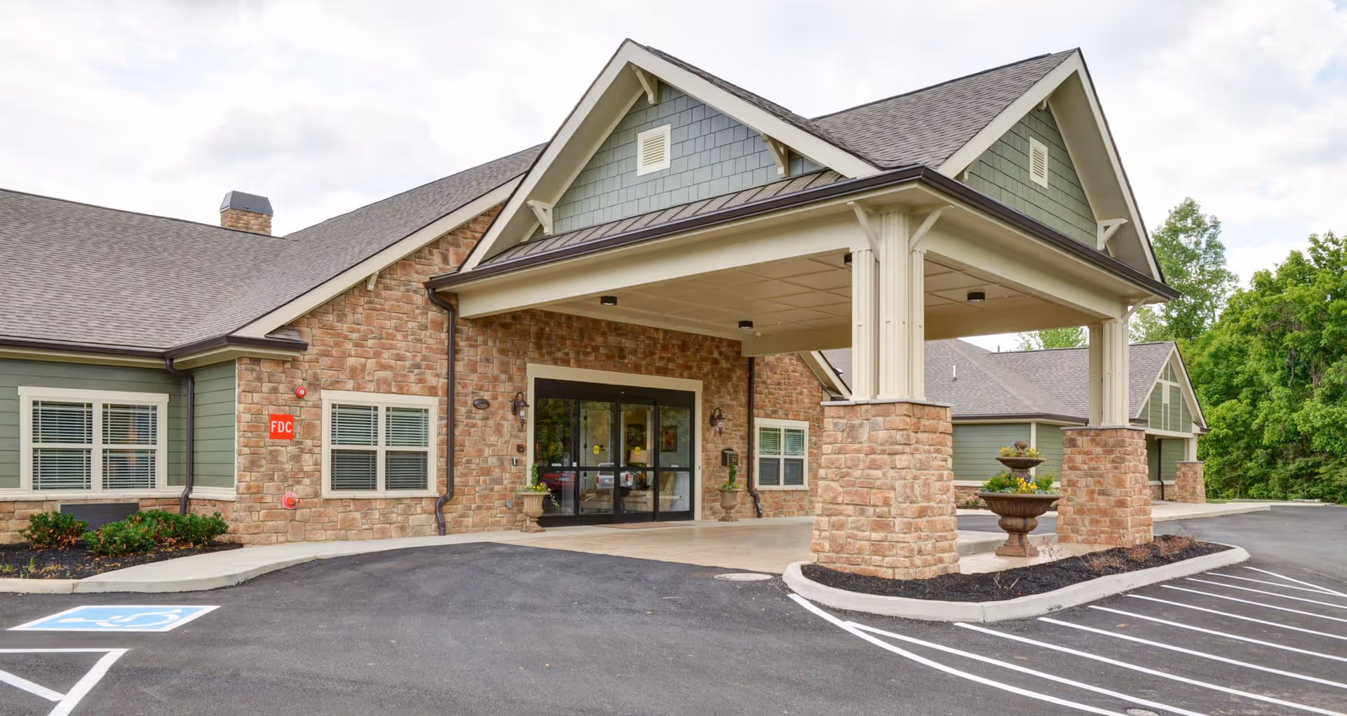 Exterior view of Dominion Senior Living of Sevierville showing the entrance with a covered drop-off area supported by stone pillars. The building features a combination of stone and green siding with multiple windows and a paved parking area including a handicapped parking space.