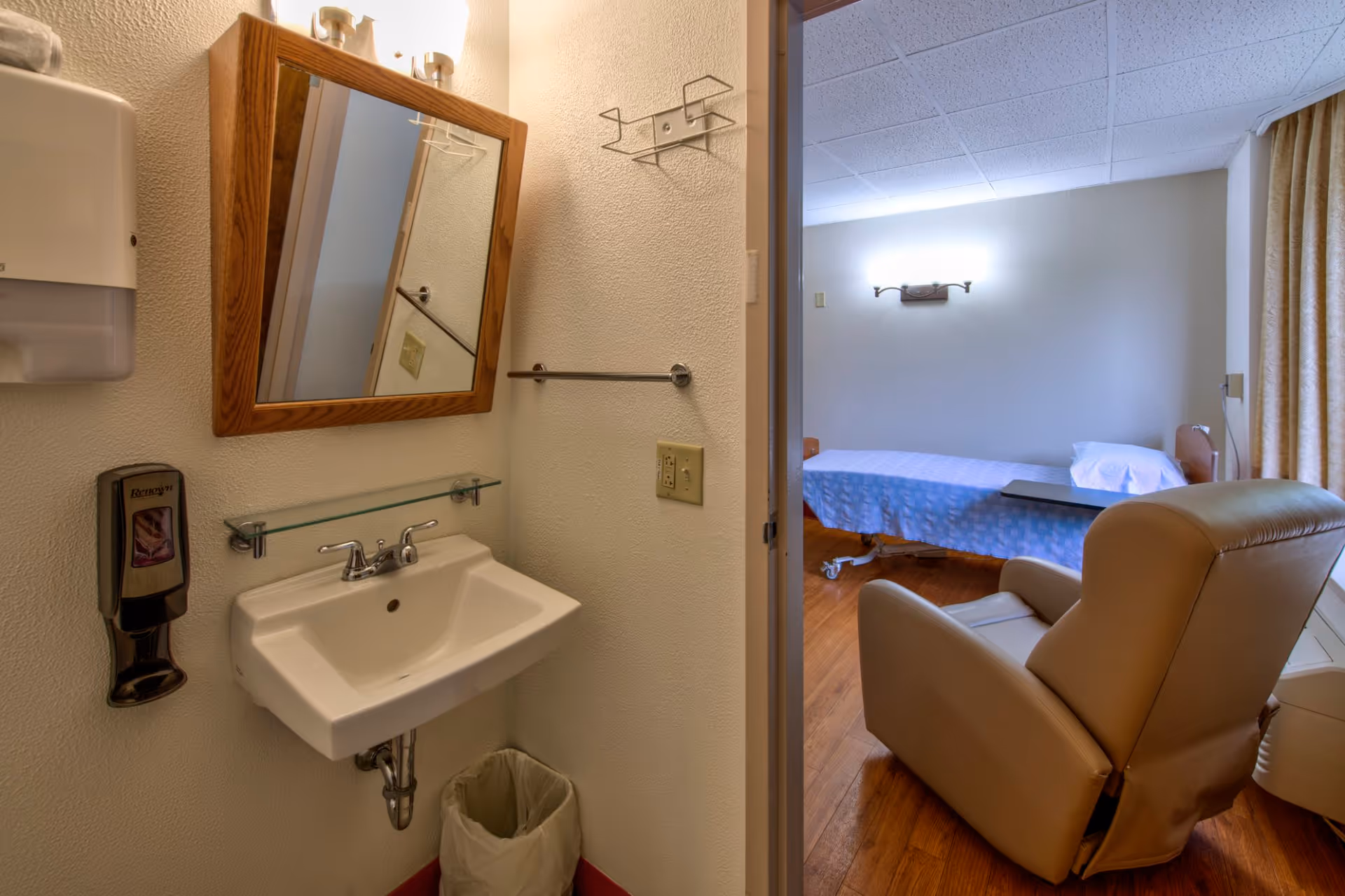 A small assisted-living bedroom showing a sink and mirror in the foreground and a bed with a recliner in the adjoining room.
