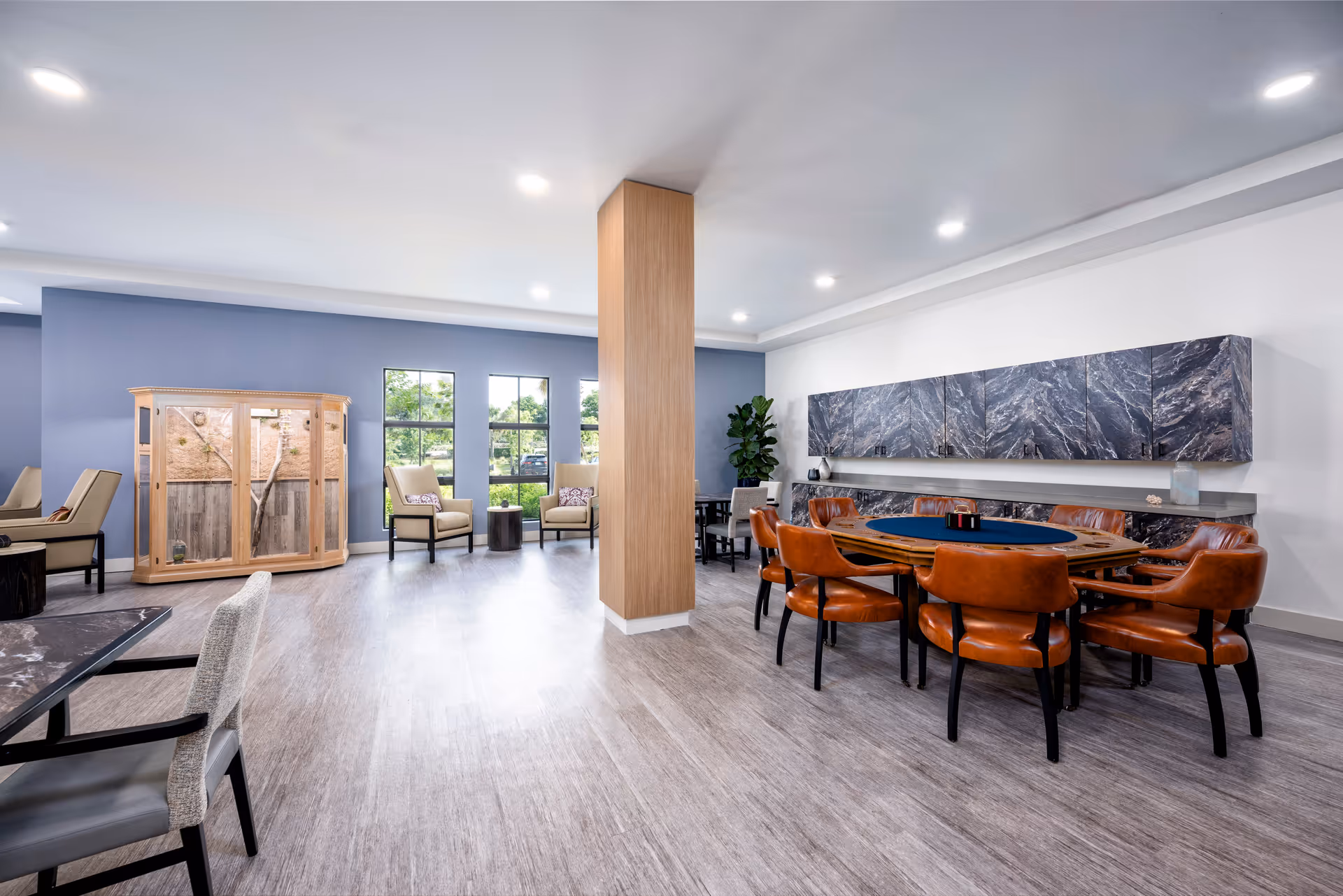 A spacious and well-lit common area in a senior living facility featuring a large wooden table with orange leather chairs, a set of beige armchairs near windows with a view of greenery outside, a wooden display cabinet, and modern cabinetry with a marble-like finish on the wall.