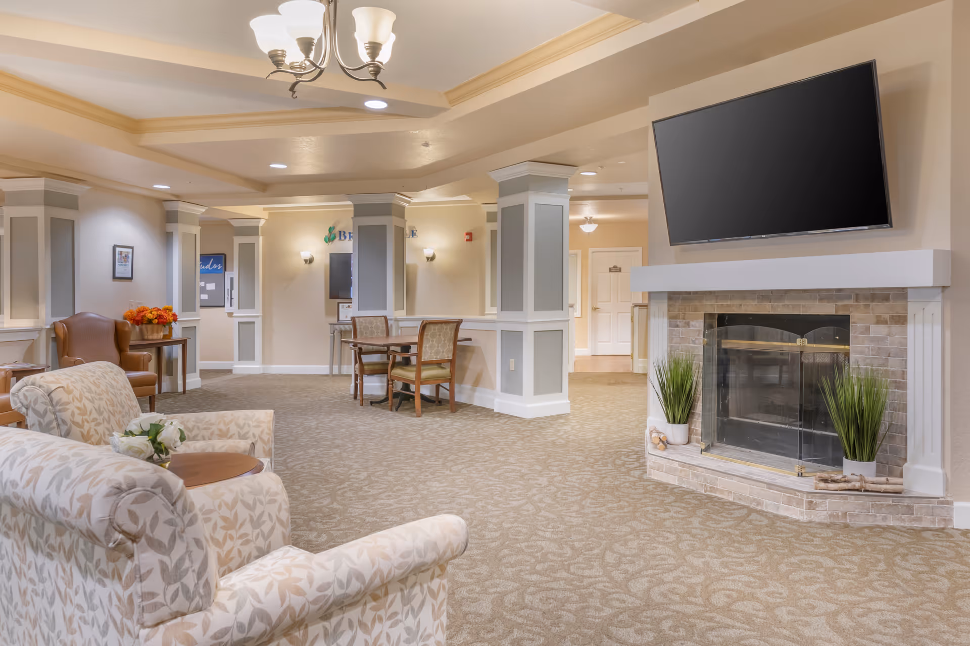 Bright senior living common lounge with patterned armchairs, a fireplace topped by a wall-mounted TV, tables and decorative columns.