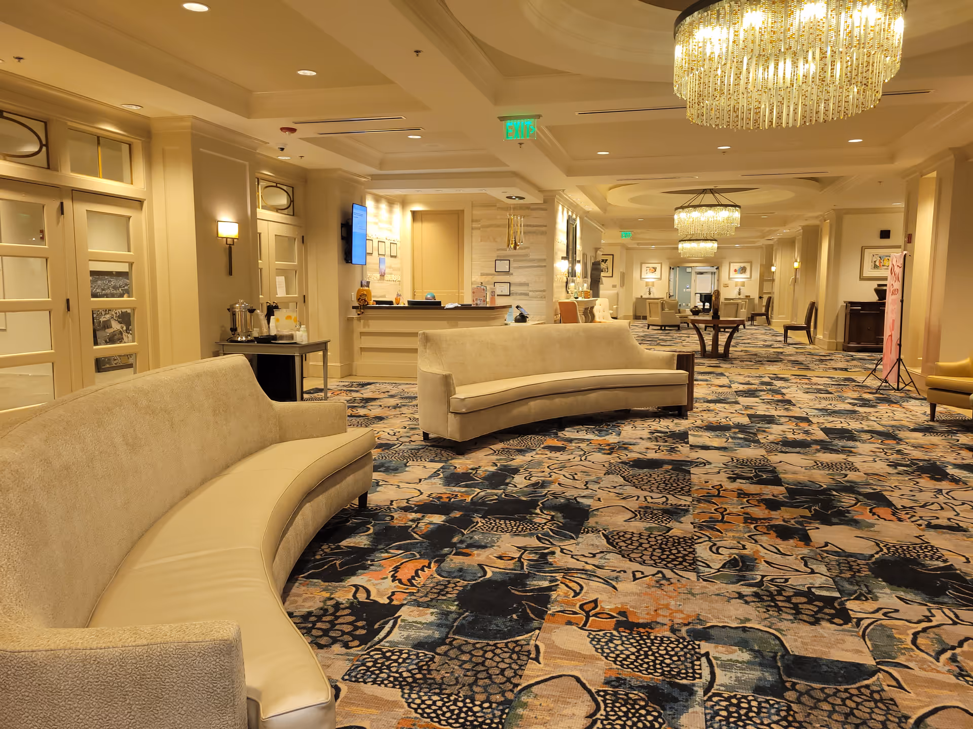 Spacious and well-lit lounge area with curved beige sofas, patterned carpet, elegant chandeliers, and a reception desk in the background. The room has cream-colored walls, framed artwork, and an exit sign on the ceiling.