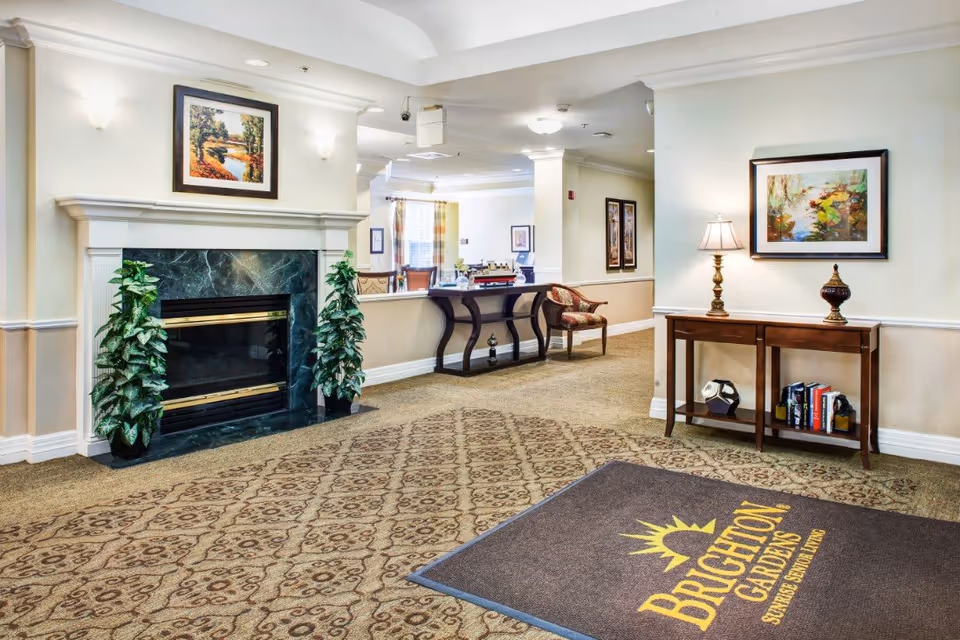 Interior view of a senior living facility lobby area with a patterned carpet, a fireplace with green marble surround, two potted plants, a wooden console table with a lamp and decorative items, framed artwork on the walls, and a mat on the floor with the text 'Brighton Gardens Sunrise Senior Living'.